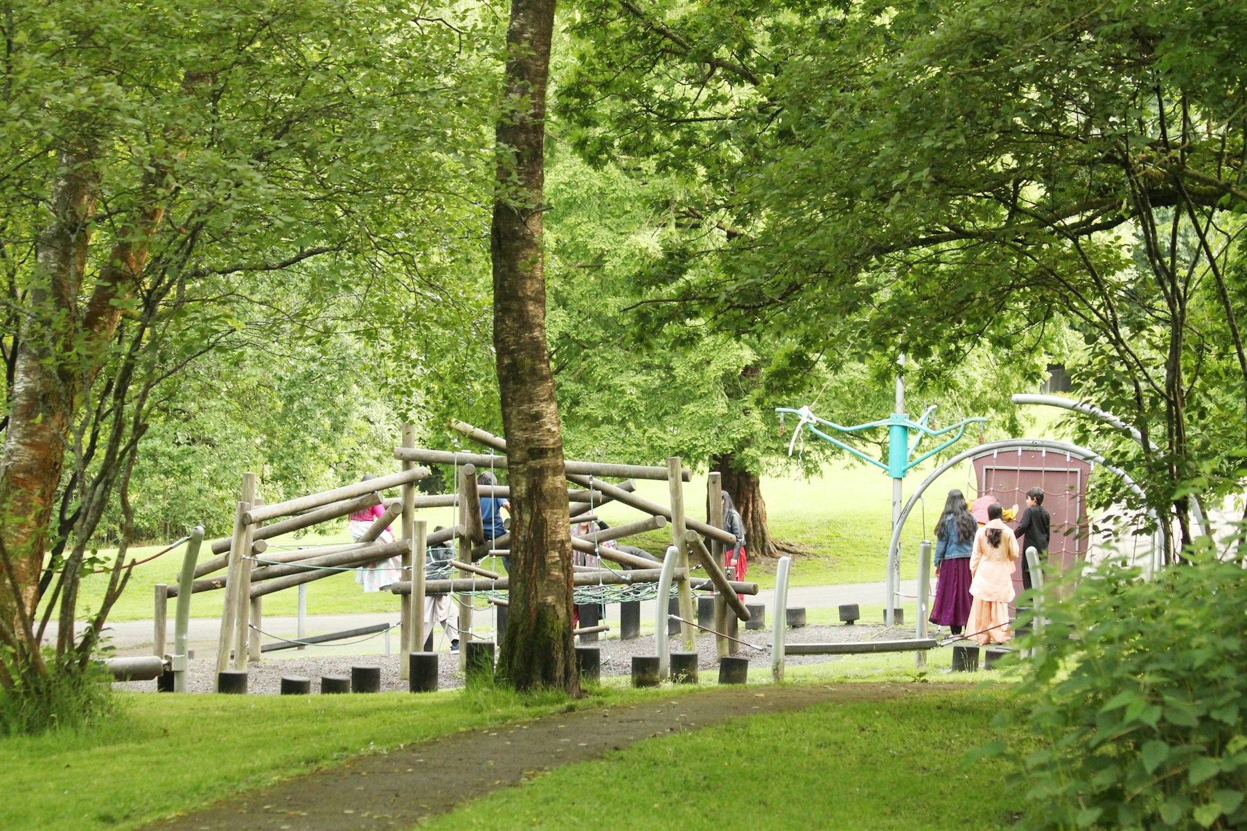 Kinderen spelen in een groene buitenruimte met speeltoestellen tussen de bomen.